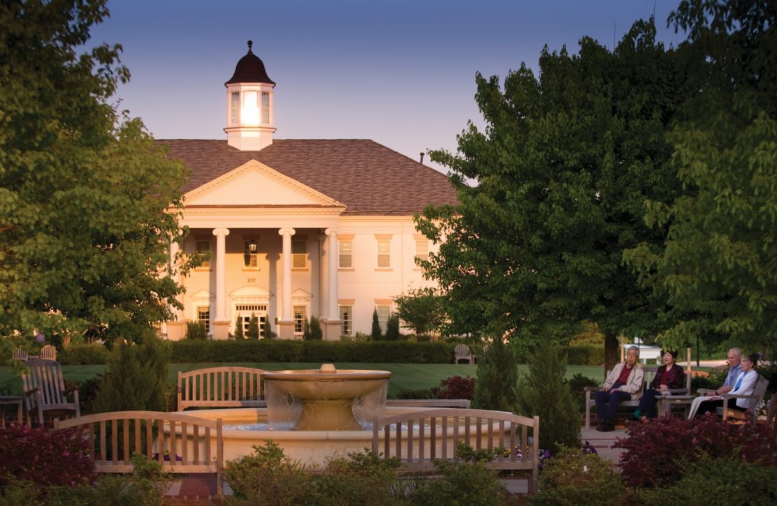 Scenic shot of The Cedars clubhouse, a retirement community in Chapel Hill NC, with a water fountain and benches in the foreground during late afternoon